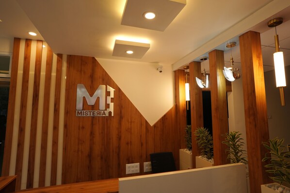 A reception area with a wooden panel wall, a large 'ME MISTERIA' sign, and modern pendant lighting.