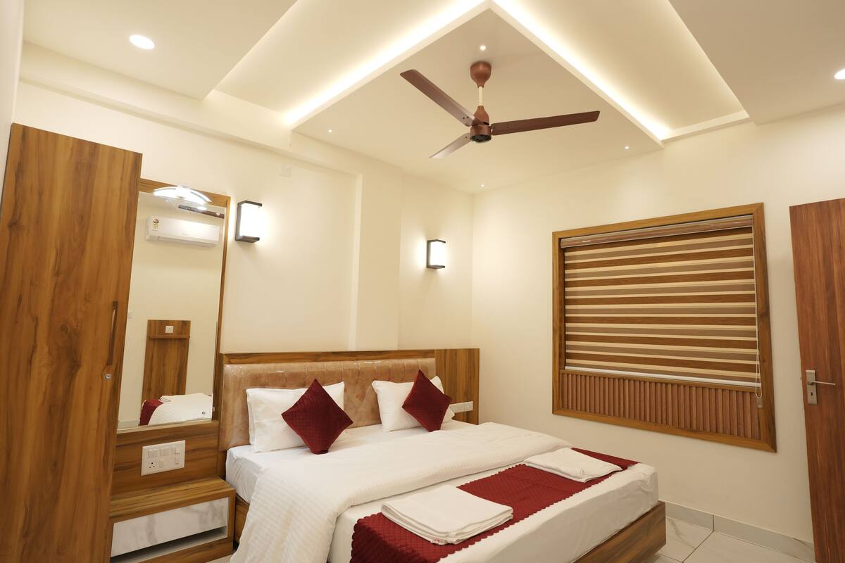 A hotel room with a wooden headboard, a ceiling fan, a large window with blinds, and a bedside table with a lamp.