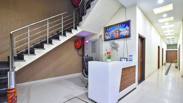 Zona de recepción de un hotel con una escalera, un mostrador de recepción, un televisor montado en la pared y un pasillo con puertas y plantas en macetas.