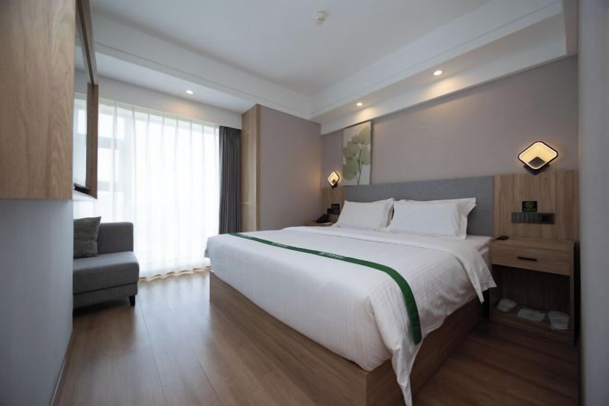 Ett modernt hotellrum med en stor säng, en grå soffa, ett träbord vid sängen och ett stort fönster med tunna, genomskinliga gardiner.