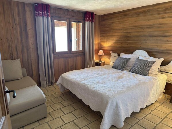 A bedroom with a bed, a chair, a window with curtains, and a wooden wall. - Chalet, 100m from ski slopes, hammam, terrace, fireplace or stove, tv, 210m², Les Saisies (Villard-sur-Doron)
