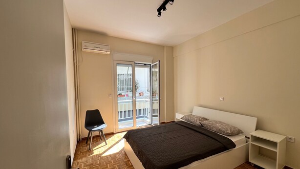 Een slaapkamer met een bed, een stoel, een klein wit nachtkastje, een plafondlamp en een schuifpui naar een balkon.
