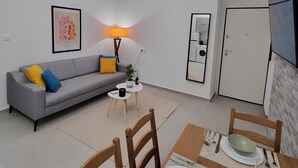 Een moderne woonkamer met een grijze bank, een klein rond tafeltje en een eetgedeelte met een houten tafel gedekt voor twee personen.