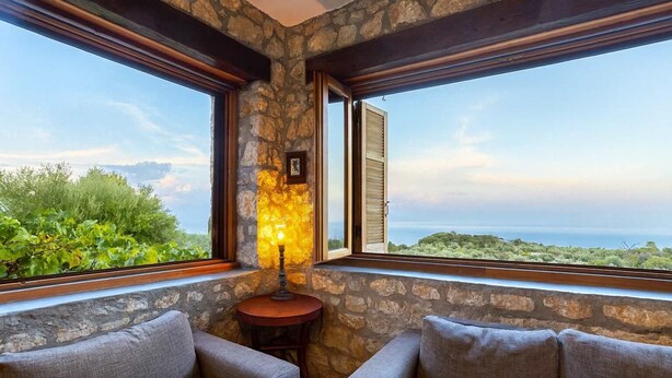 A cozy room with stone walls, two grey sofas, a small round table with a lamp, and large windows offering a scenic view of greenery and the sea. - Stone Farmhouse in Groves - Sea & Mountain Views (Achladochori)