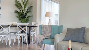 A living room with a teal armchair, a beige sofa, a dining table with white chairs, a potted plant, and a lamp. - Horseshoe View 1A Scrymgour Rd Port Elliot (Port Elliot)