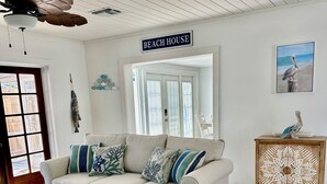Una sala de estar con un sofá beige, una mesa de centro blanca, un mueble de madera con un diseño decorativo, un ventilador de techo y un letrero que dice "BEACH HOUSE".