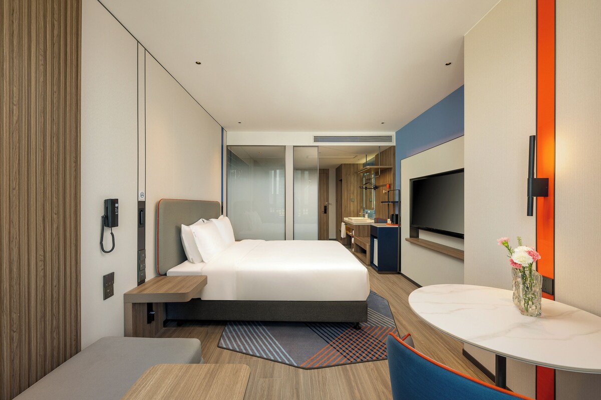 Una habitación de hotel moderna con una cama grande, una mesita de noche y un pequeño escritorio con una silla. Hay un televisor montado en la pared y un florero con flores sobre una mesa.