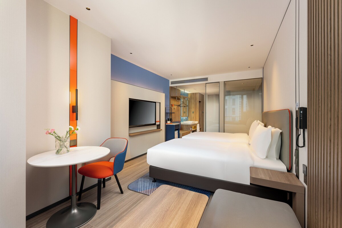 Habitación de hotel moderna con una cama grande, una mesita con una silla, televisor y baño visible a través de un separador de vidrio.
