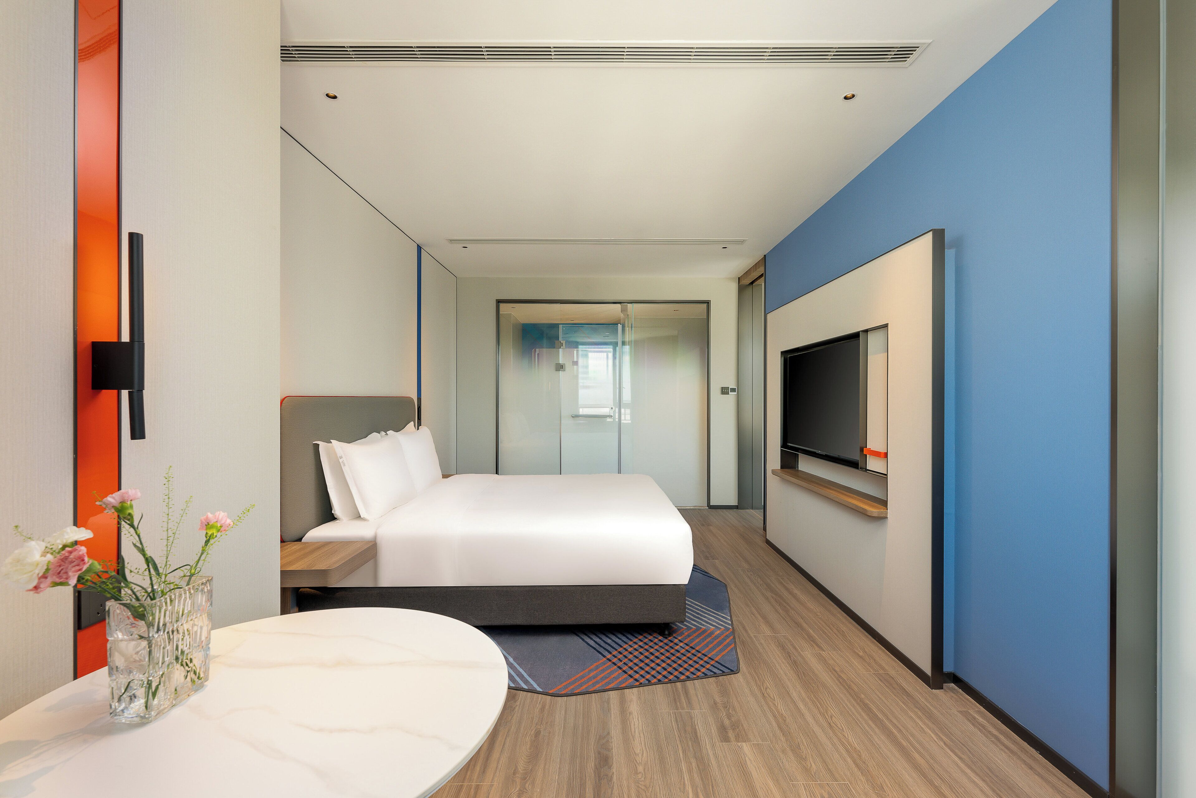 Una habitación de hotel moderna con una cama grande, un televisor de pantalla plana, un florero con flores y una mesa redonda.