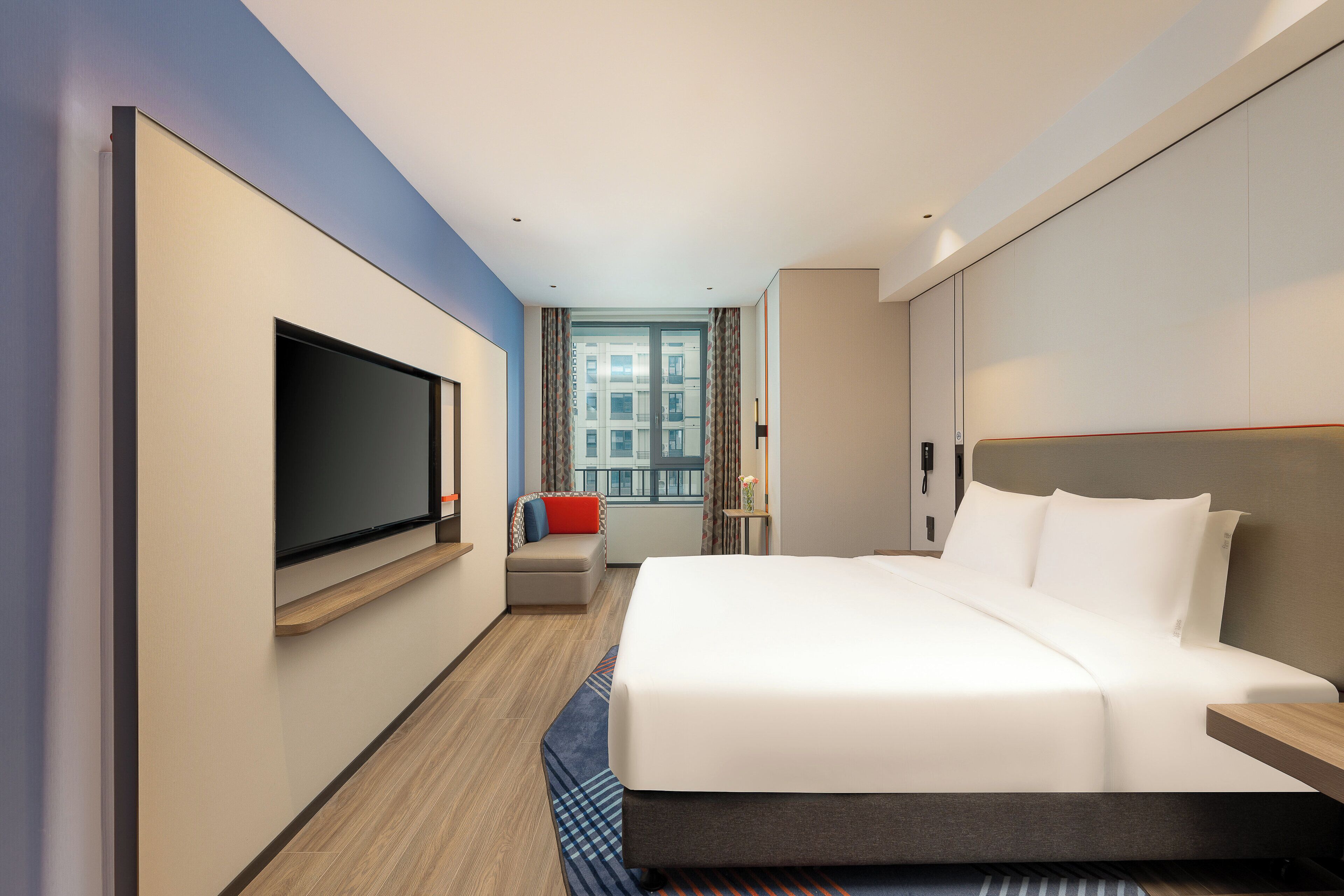 Una habitación de hotel moderna con una cama grande, un televisor de pantalla plana montado en la pared, una zona de estar con un cojín rojo y una ventana con vistas a la ciudad.