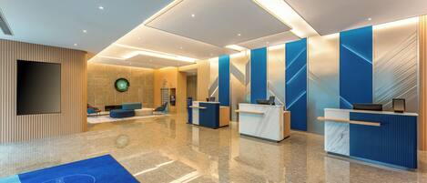A modern hotel lobby with a reception desk, seating area, and a large screen.