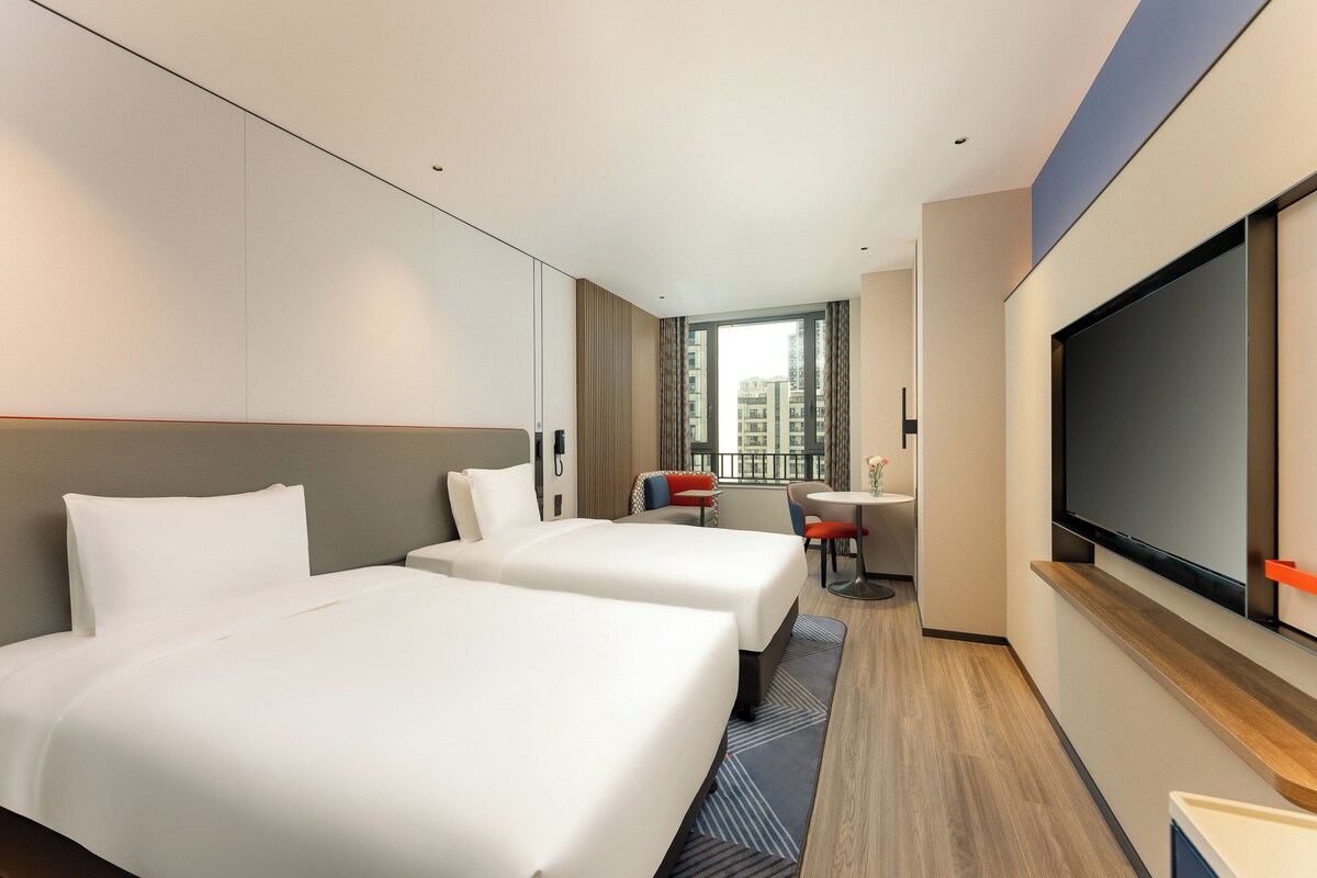 A hotel room with two beds, a TV, a desk, and a window with a city view.
