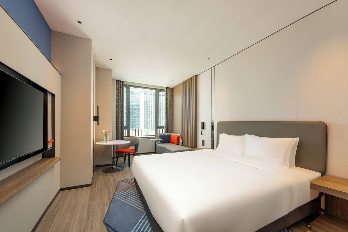 A modern hotel room with a large bed, a flat-screen TV, a small table with chairs, and a window with a city view.