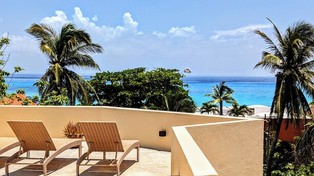 Un balcon doté de fauteuils en osier, donnant sur une plage bordée de palmiers et offrant une vue sur un ciel dégagé.