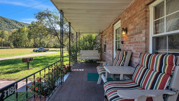 A covered porch with outdoor seating, a brick wall, and a view of a road and greenery.