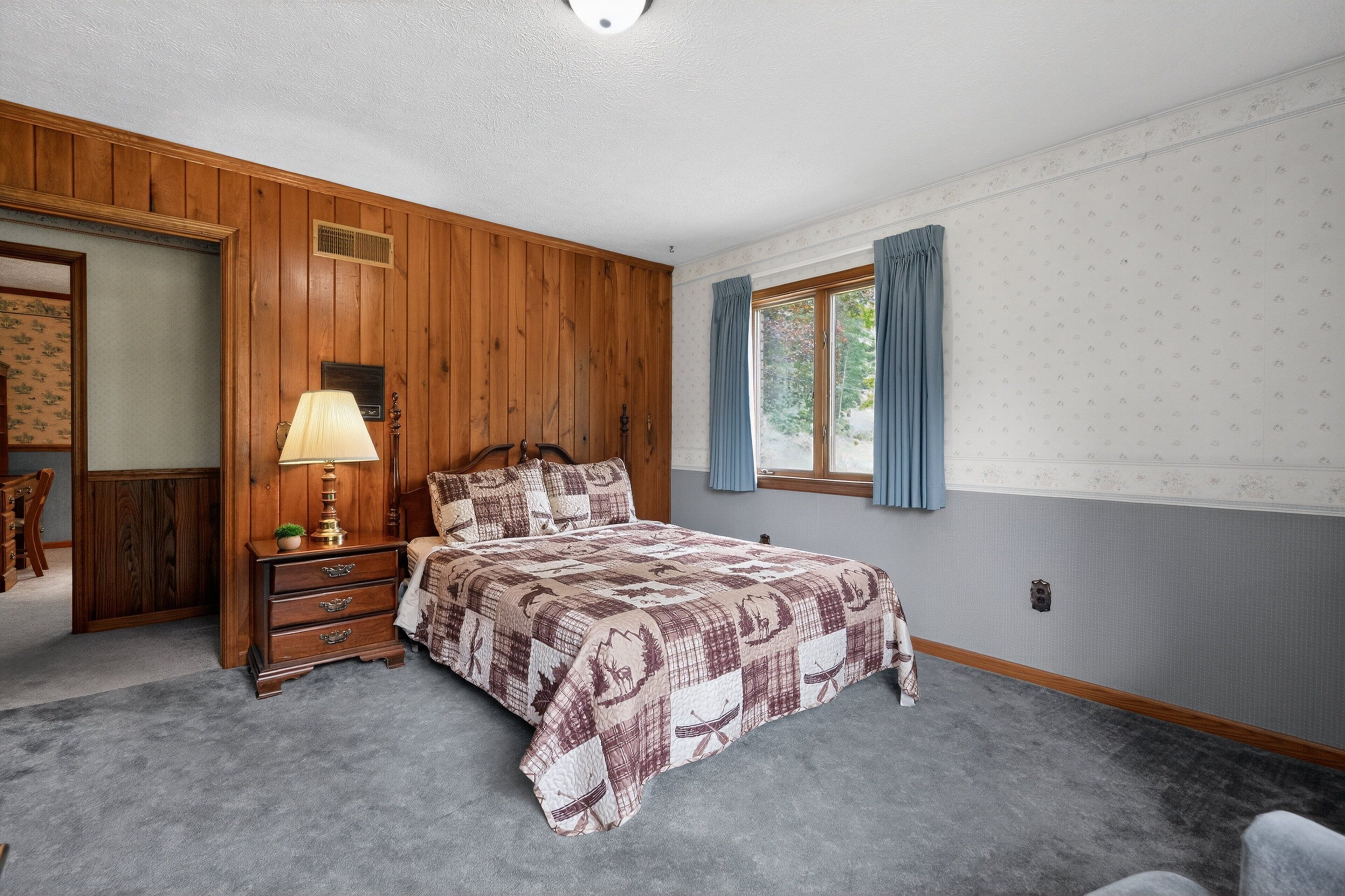 A bedroom with a wooden paneled wall, a bed with a patterned bedspread, a nightstand with a lamp, and a window with curtains.