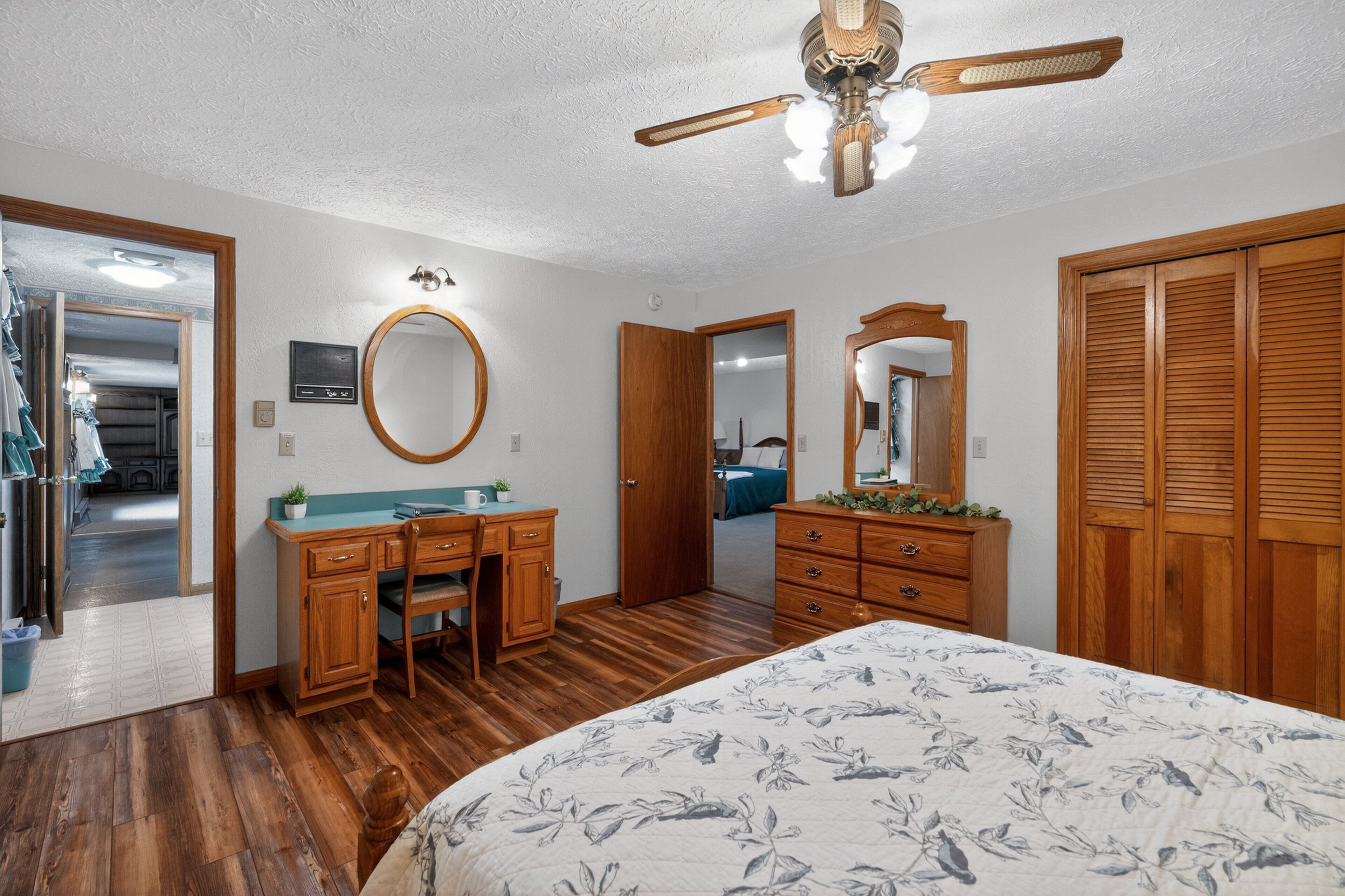 A bedroom with a wooden bed, a desk with a chair, a dresser, a ceiling fan, and a large mirror.