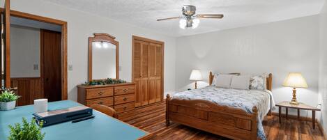 Una camera da letto con un letto di legno, una scrivania con un computer, un comò, uno specchio e una ventola a soffitto.