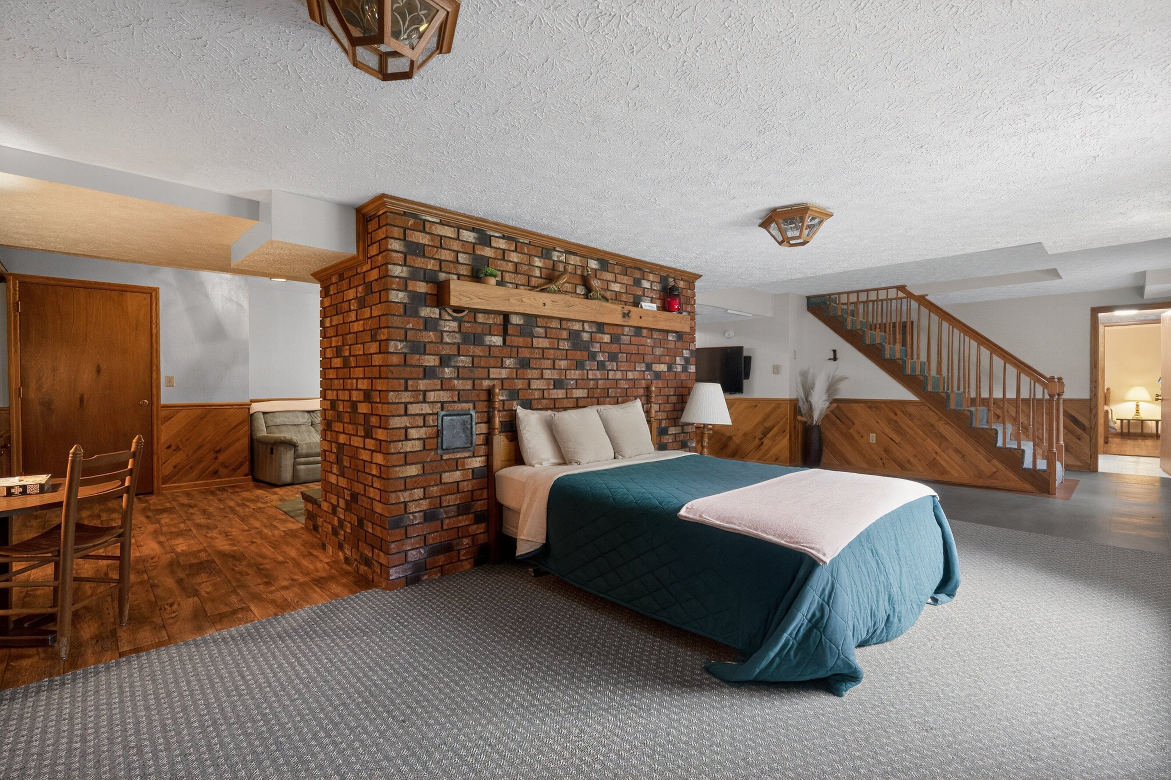 A bedroom with a brick accent wall, a bed with a teal bedspread, a wooden chair, and a staircase.