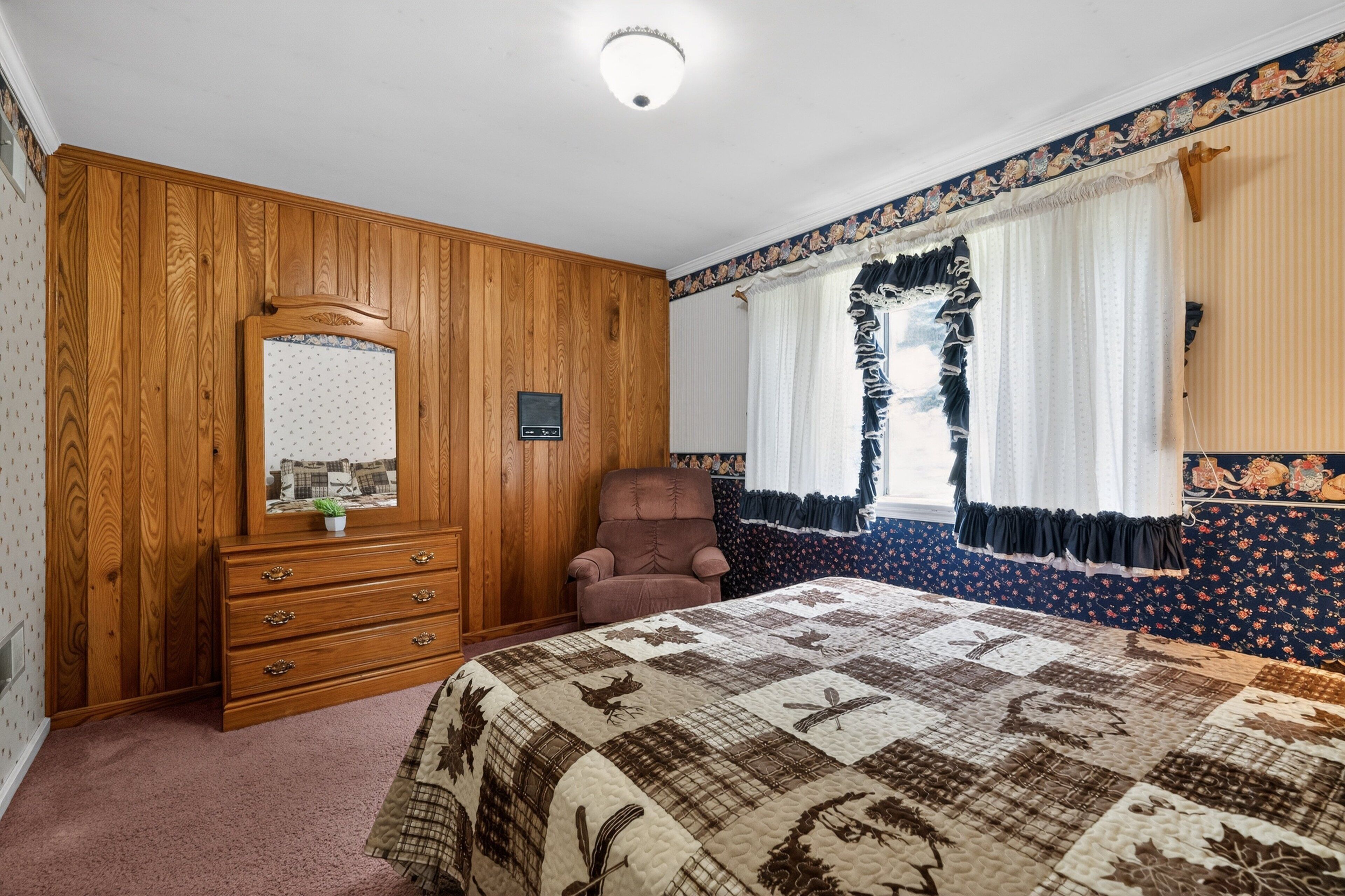 A bedroom with a wooden dresser, a chair, a bed with a quilt, and a window with curtains.