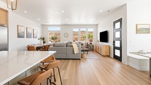 Een ruime woonkamer met een grote bank, een eettafel met barstoelen, en een keuken met een wit aanrecht en houten kastjes.