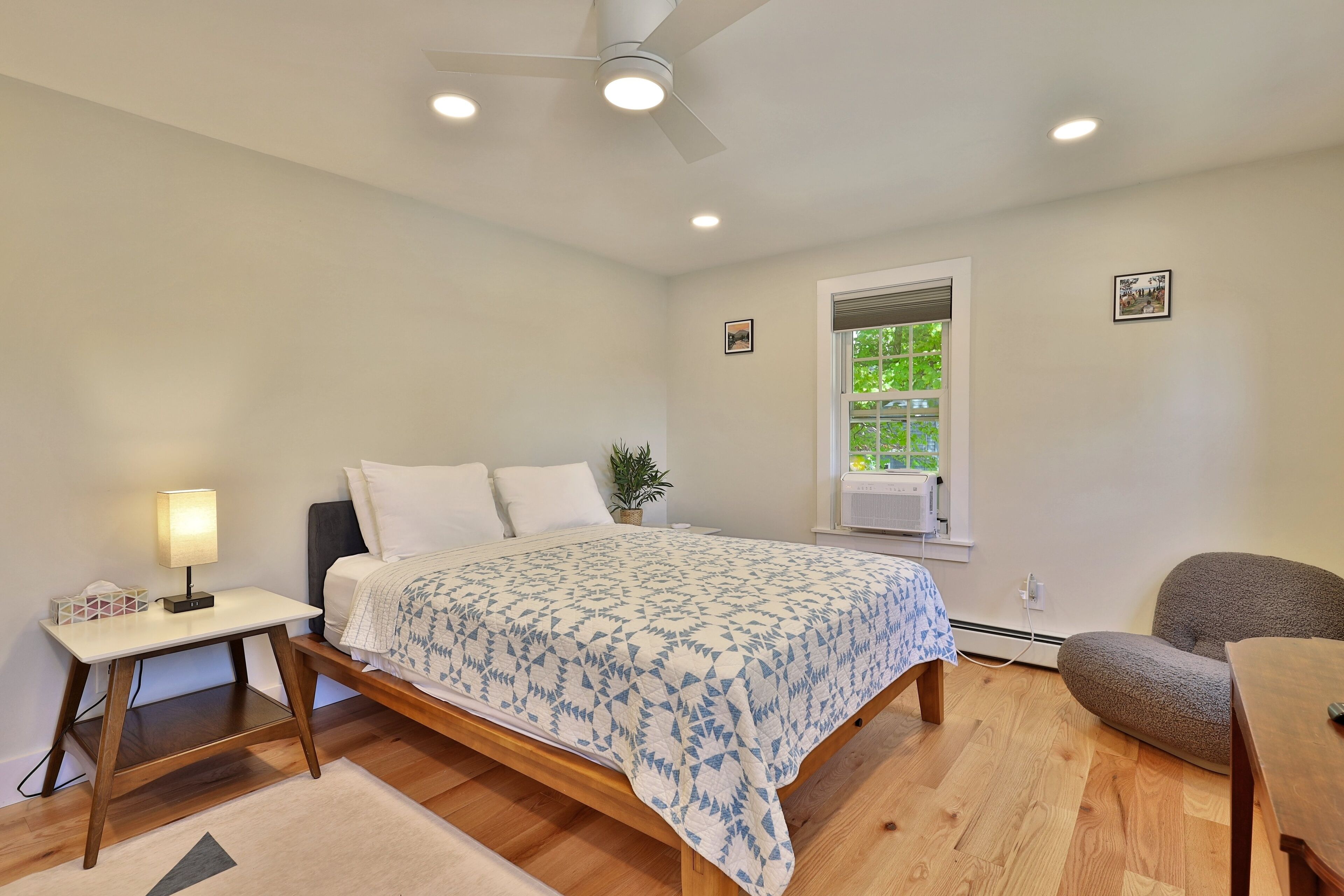 A bedroom with a bed, bedside table, lamp, chair, and a window with a view of greenery.