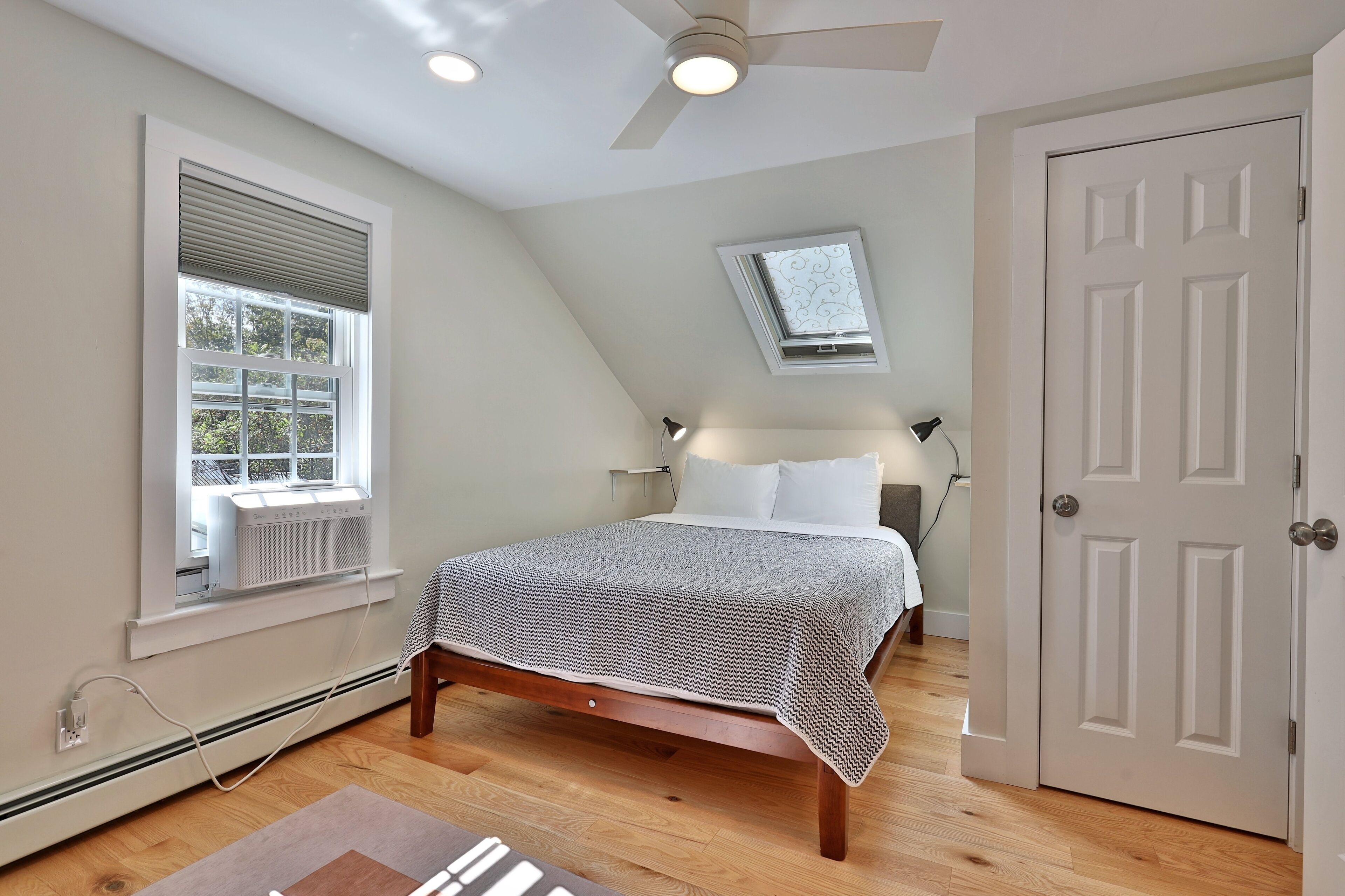 Een slaapkamer met een bed, een plafondventilator, een raam met zonwering, een deur en een klein dakraam.