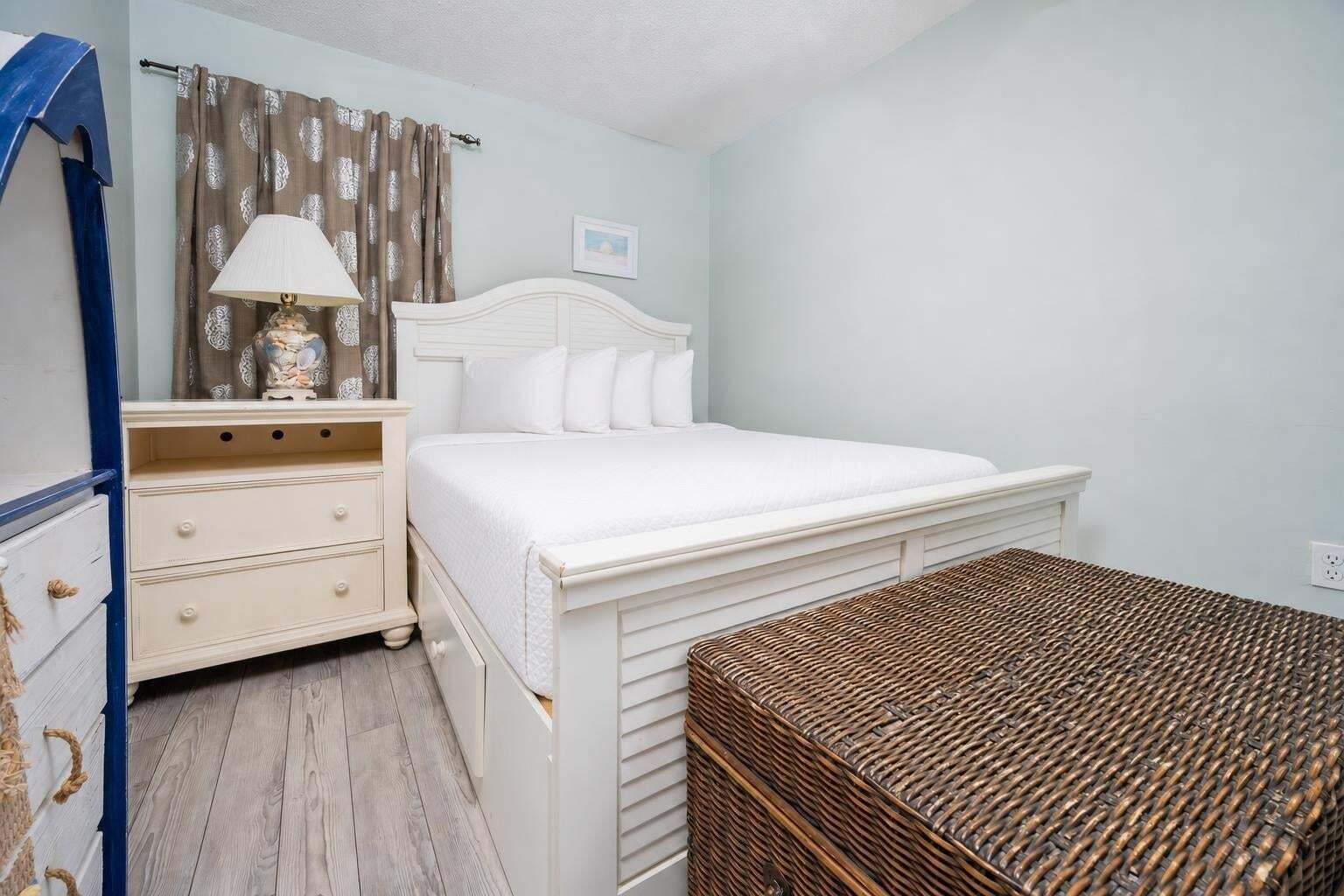 A bedroom with a bed, a nightstand, a lamp, a picture on the wall, and a wicker bench.