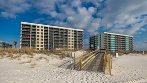 Un hotel frente a la playa con varios pisos, una playa arenosa en primer plano y cielo despejado.