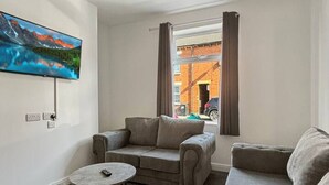 Uma sala de estar moderna com um sofá, um pufe, uma mesa de centro redonda e uma televisão de tela plana na parede.