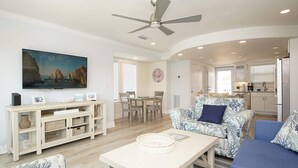 A living room with a ceiling fan, a flat-screen TV, a dining area with a table and chairs, and a kitchen in the background.