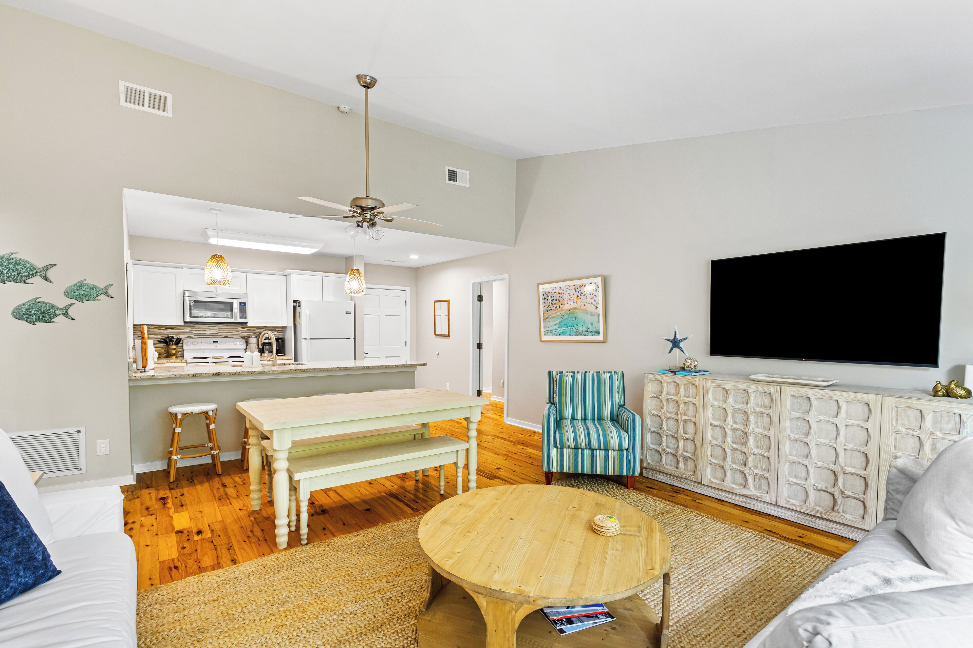 A living room with a wooden coffee table, a striped armchair, a white sofa, and a kitchen area with a microwave and cabinets.