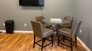 A conference room with a round glass table, six leather chairs, a black trash bin, and a mounted flat-screen TV on the wall.