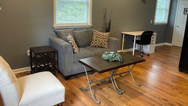 A living room with a grey sofa, a white armchair, a wooden coffee table, and a small side table. There is a window with blinds, a door, and a small desk with a chair.
