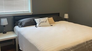A neatly made bed with a white comforter and a pillow with "BE OUR GUEST" text, a bedside lamp, and a window with blinds.