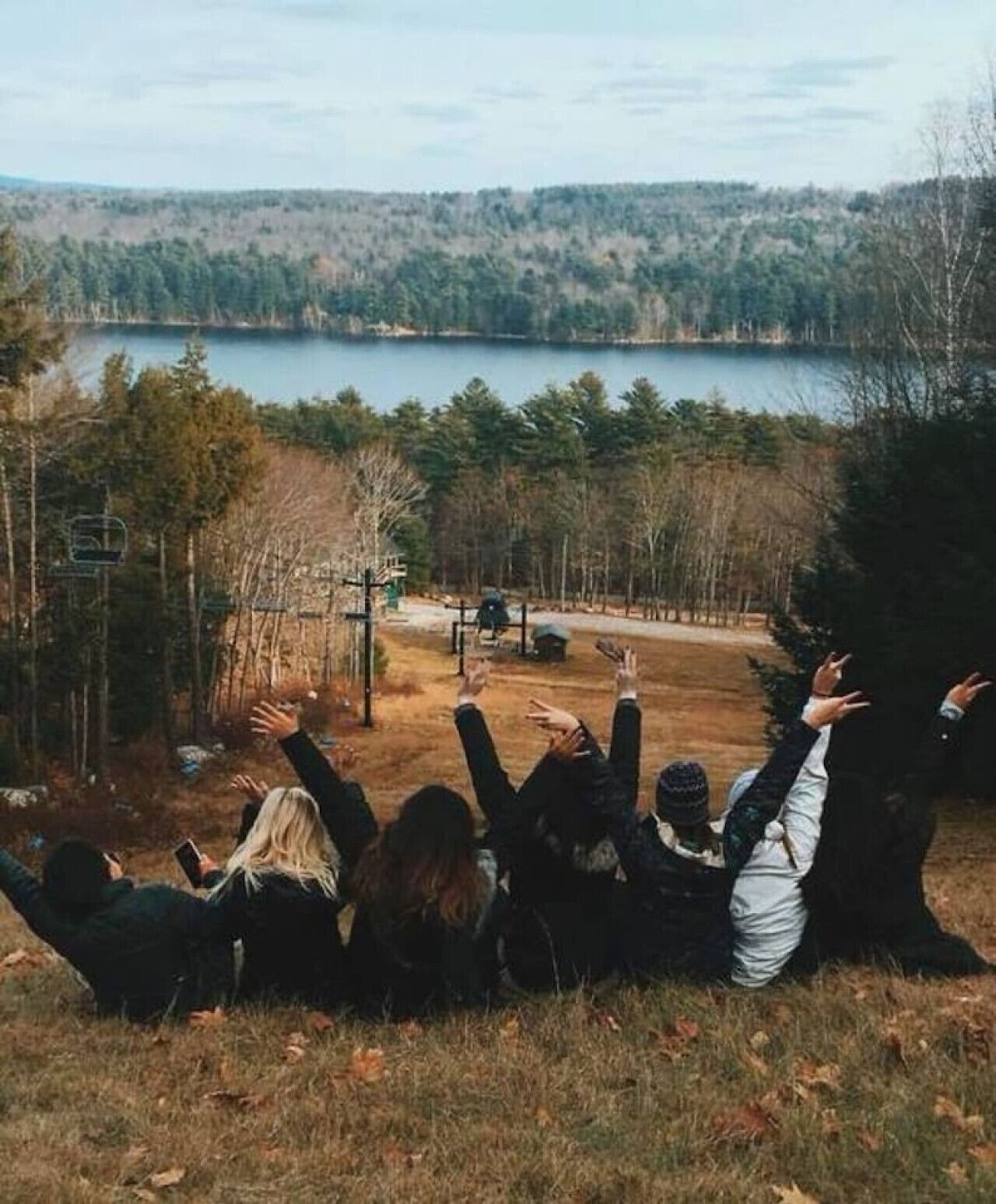 Eine Gruppe von Menschen sitzt auf einem grasbewachsenen Hügel mit Blick auf einen See und einen bewaldeten Bereich.