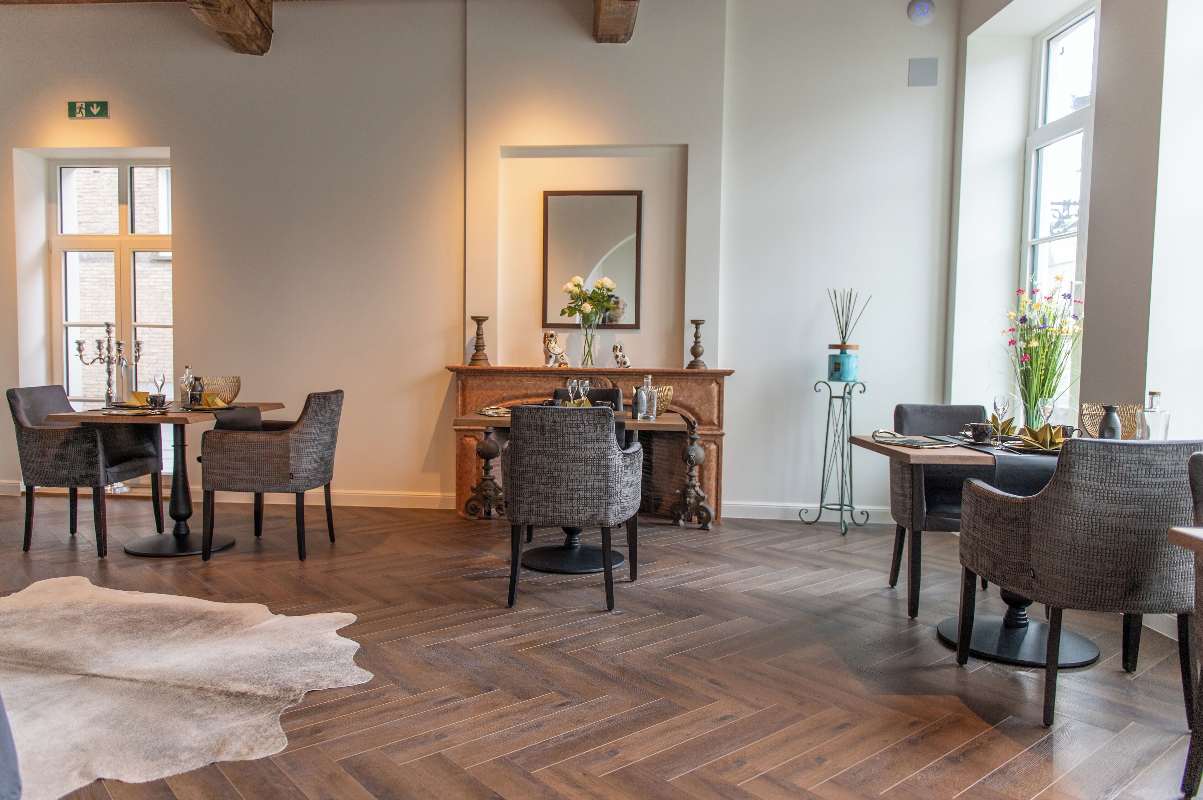 Un coin repas avec un parquet en bois disposé en chevrons, une cheminée et un miroir au-dessus. Quatre chaises sont disposées autour d’une table recouverte d’une nappe blanche.