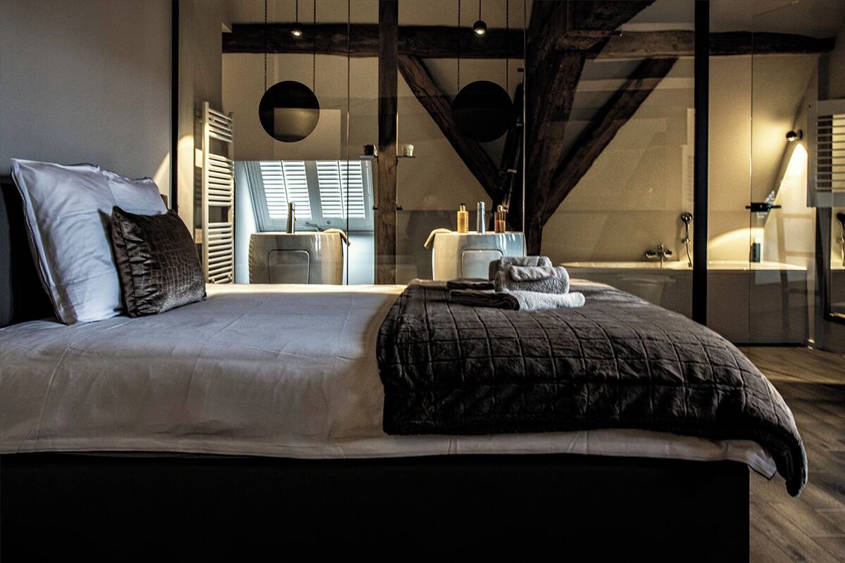 A modern bedroom with a large bed, a small seating area, and a bathroom visible in the background.
