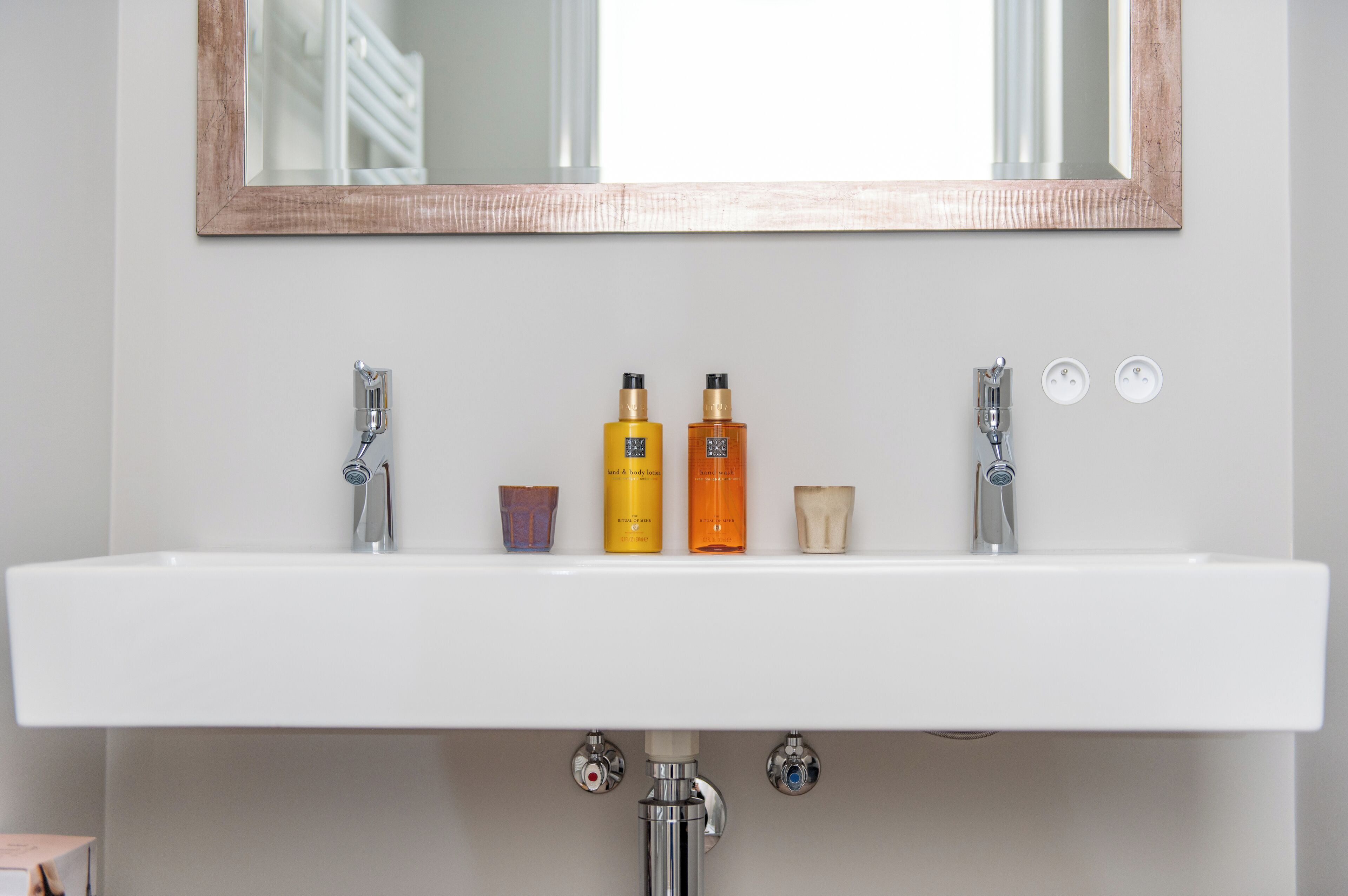Un lavabo blanc dans une salle de bain, avec deux robinets, un miroir au-dessus et trois flacons de lotion posés sur le bord du lavabo.