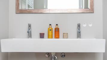 Un lavabo blanc dans une salle de bain, avec deux robinets, un miroir au-dessus et trois flacons de lotion posés sur le bord du lavabo.