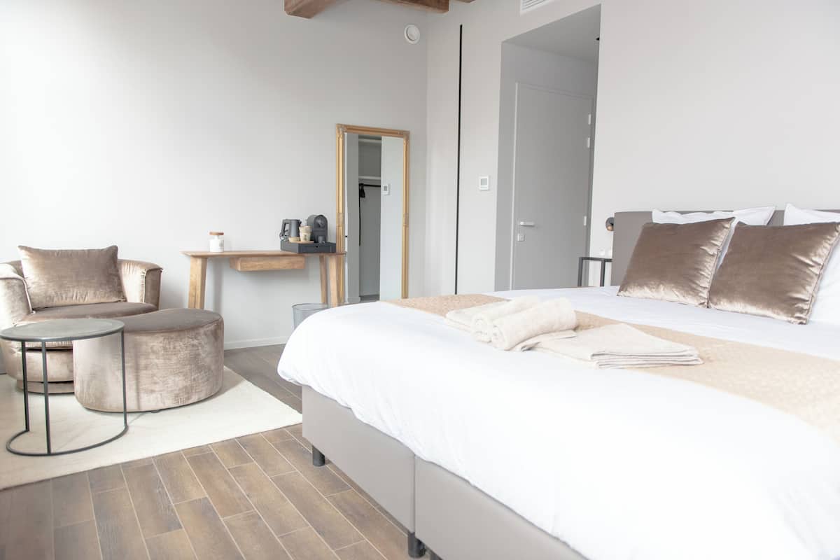 A modern bedroom with a large bed, two bedside tables, a round ottoman, a small table, and a mirror.