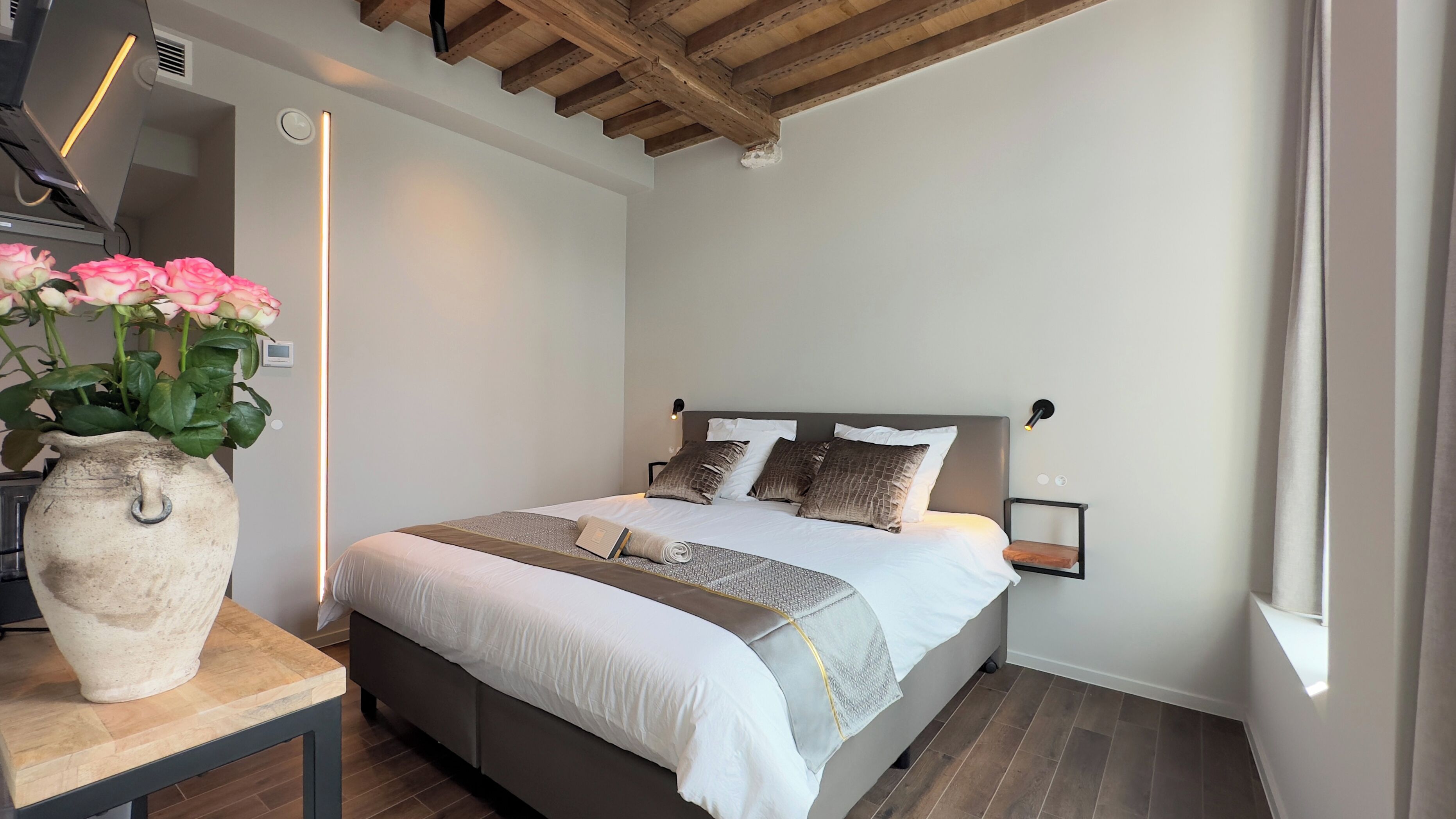 A bedroom with a bed, bedside tables, a vase with flowers, and a wooden ceiling.