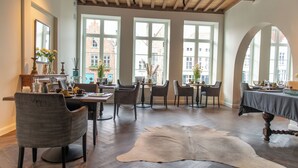 Un comedor con suelo de madera, varias mesas y sillas, y grandes ventanales que ofrecen vistas a edificios.