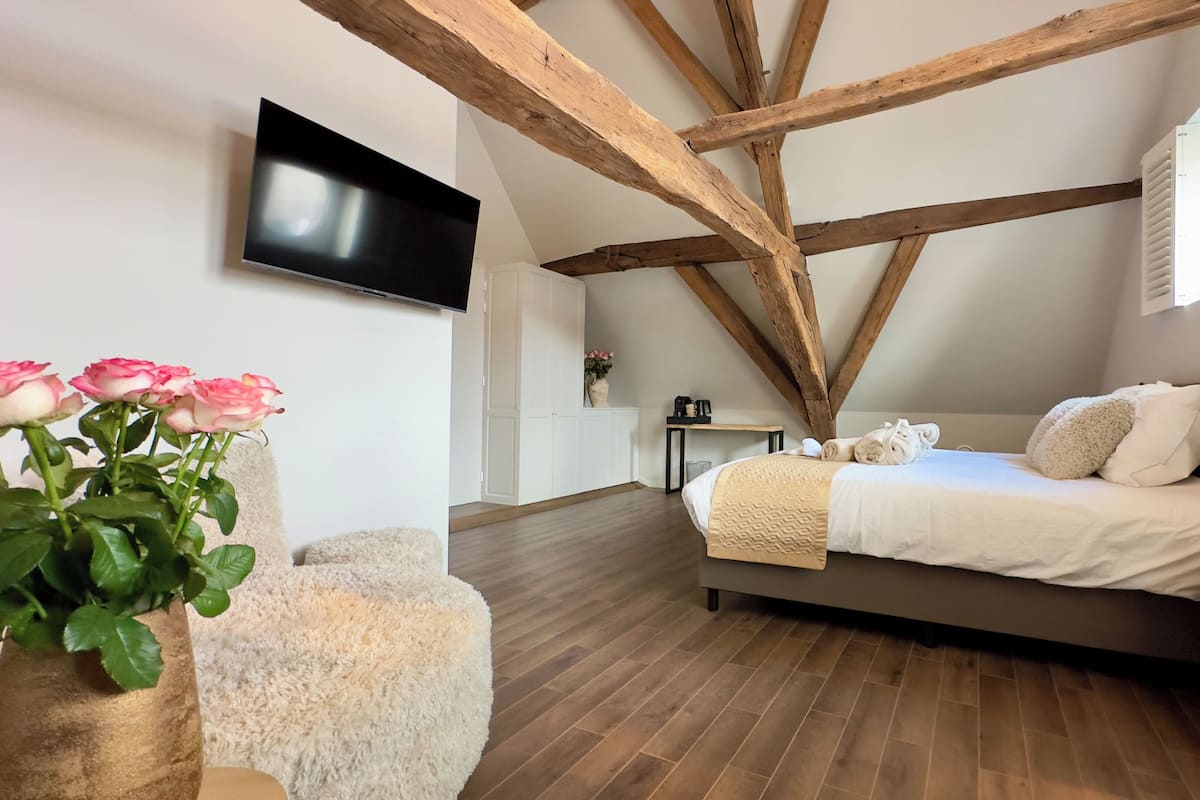 A bedroom with a wooden ceiling, a bed with white bedding, a TV mounted on the wall, and a fluffy chair.