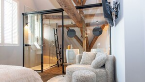 Habitación moderna y bien iluminada con puerta de vidrio que da a la zona de ducha, un rincón de descanso con cojines mullidos y un televisor montado en la pared.