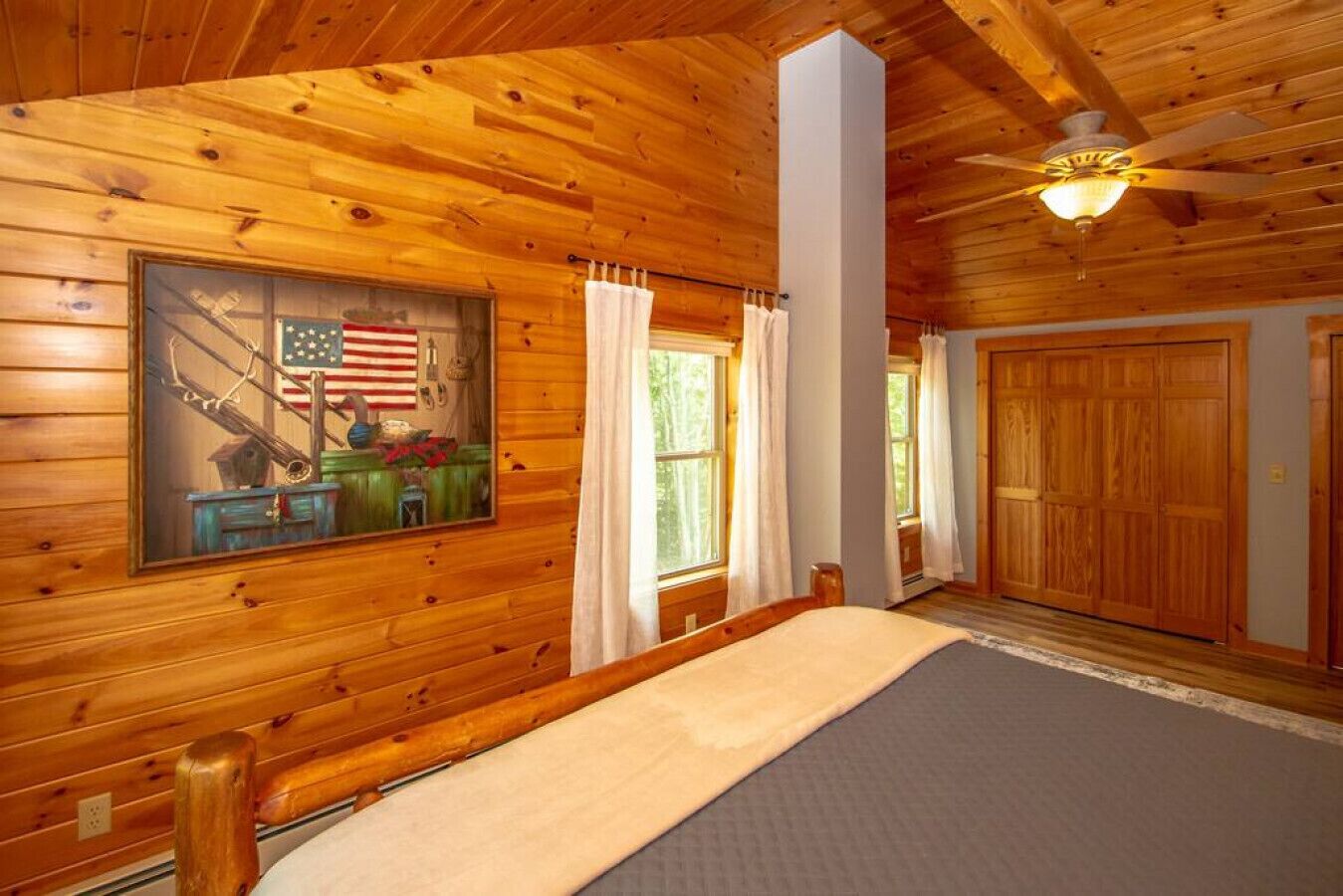 A log cabin bedroom with a bed, a ceiling fan, and a large window with curtains.