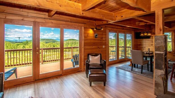 A spacious room with wooden floors, a large window with a scenic view, a leather chair, and a dining area with a table and chairs. - Spacious Log Home Overlooking 40 White Mnt Peaks with Extreme Privacy: Host Weddings (Brownfield)