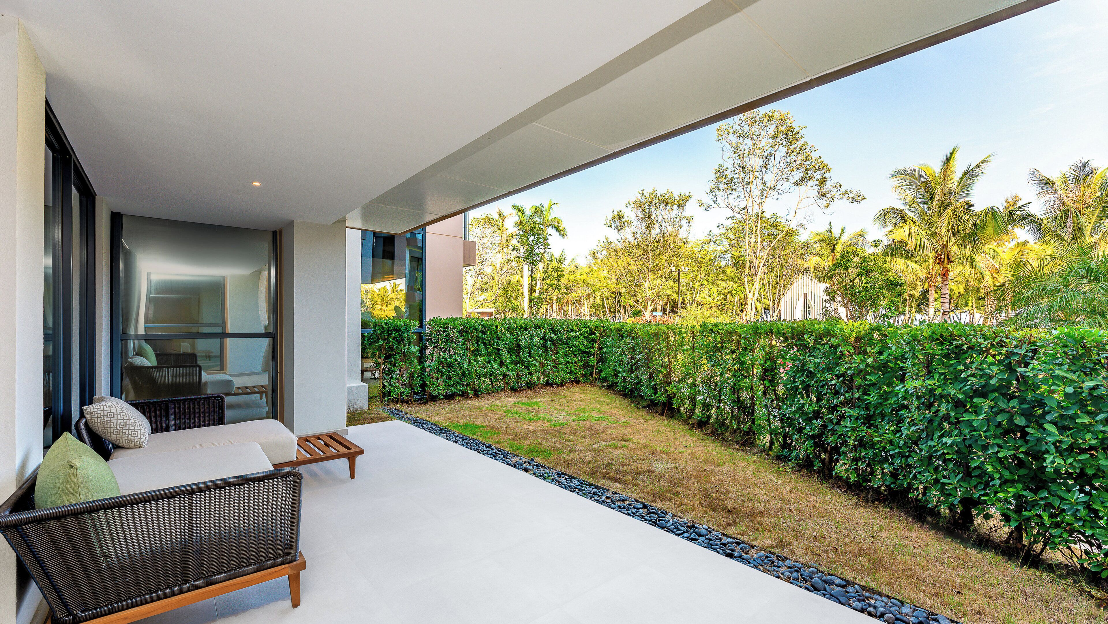 A modern outdoor seating area with a sofa and a chair, a well-maintained hedge, and a view of a lush garden with trees and a white building in the background.