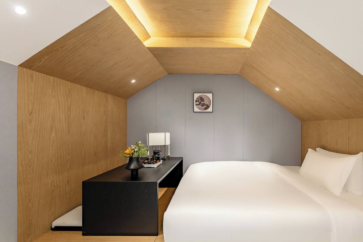 Un dormitorio moderno con techo inclinado, una cama, una mesita de noche con lámpara y una pequeña obra de arte en la pared.