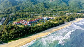 Luftaufnahme eines Küstenresorts mit mehreren Gebäuden, einem Strand und einem bewaldeten Gebiet.